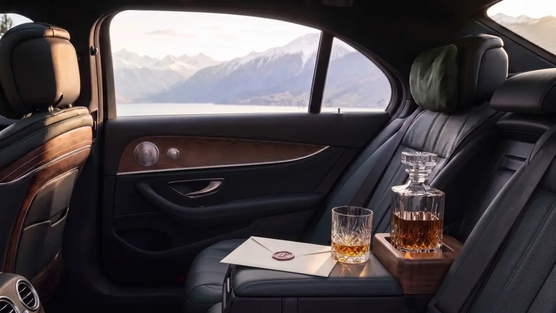 Fond einer Limousine mit Leder und Holz, auf dem Sitz ein mit Wachs versiegelter Umschlag, eine Whiskykaraffe und ein Glas, draußen ein See und Berge
