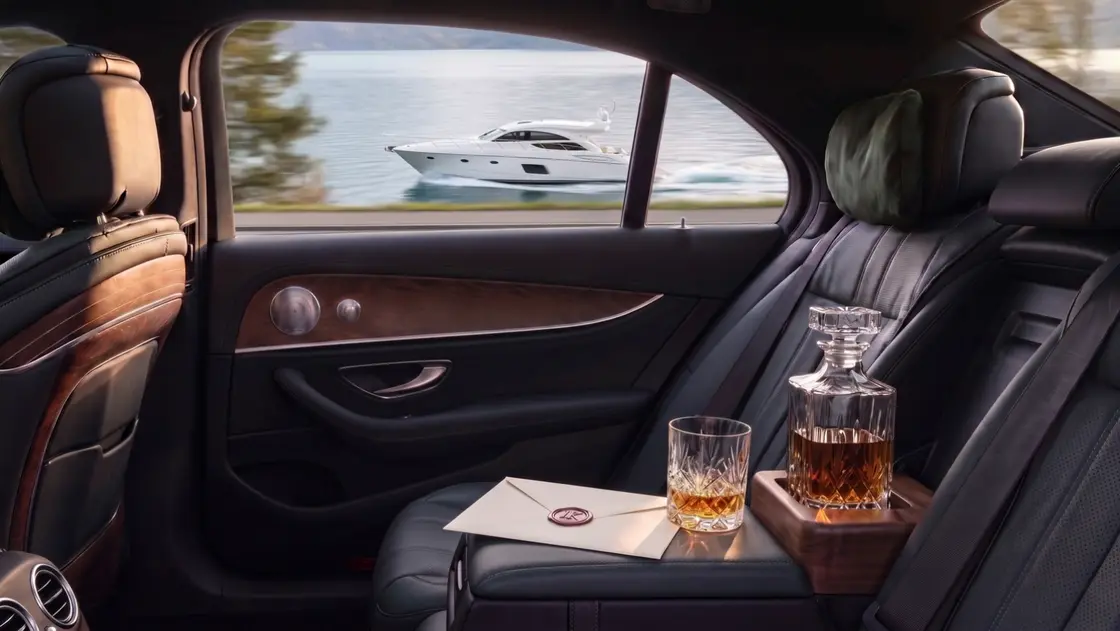 Fond einer Limousine mit Leder und Holz, darauf eine Whiskykaraffe und ein Glas, durch das Fenster eine weiße Motoryacht auf einem See